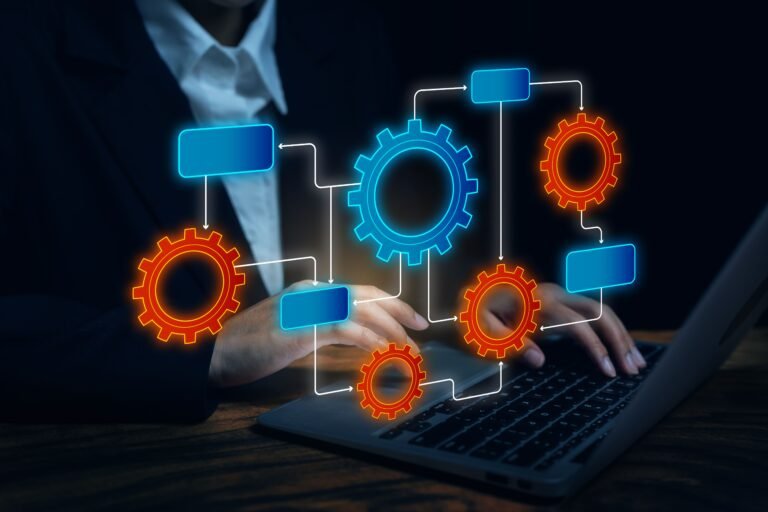 Person in a dark suit typing on a laptop, with glowing blue and orange gears connected by white arrows to blue boxes in a flowchart-like diagram.
