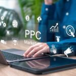 Blue-shirted person typing on a laptop with floating PPC marketing icons—graph, globe, megaphone, target, and dollar symbols—around the keyboard.