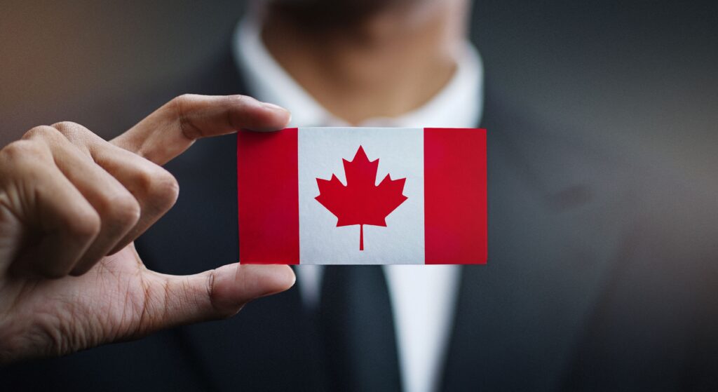 Hand holding a card with the Canadian flag: red side bars and white center with a red maple leaf, against a dark suit.