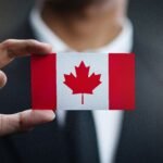 Hand holding a card with the Canadian flag: red side bars and white center with a red maple leaf, against a dark suit.