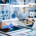 Hands hover over a tablet and laptop with floating analytics dashboards showing pie, bar and line charts in blue, orange and teal.