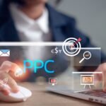Hands on a laptop with floating PPC interface icons: blue PPC text, target bullseye, envelope, chart, magnifier, ad window, and currency symbols.