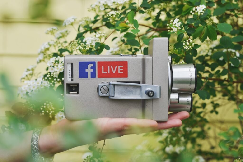 Grey vintage camera with a Facebook Live sticker, held in a hand among green leaves and white flowers, two chrome lenses visible.