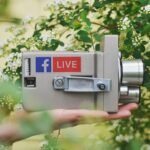 Grey vintage camera with a Facebook Live sticker, held in a hand among green leaves and white flowers, two chrome lenses visible.