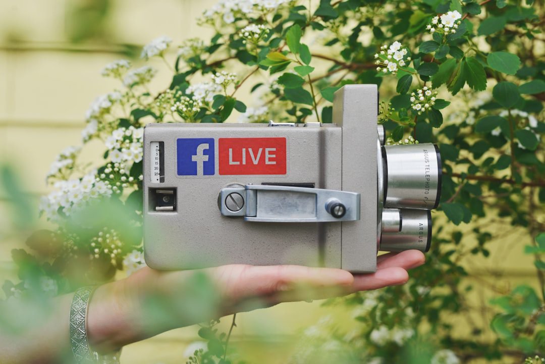 Grey vintage camera with a Facebook Live sticker, held in a hand among green leaves and white flowers, two chrome lenses visible.